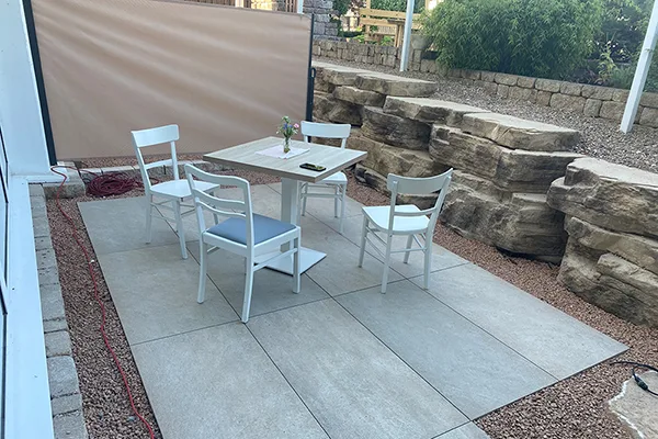 Ein weißer Esstisch mit vier Stühlen auf einer Terrasse mit beheizbaren Fliesen. Im Hintergrund sind eine natürliche Steinmauer und eine beige Markise.
