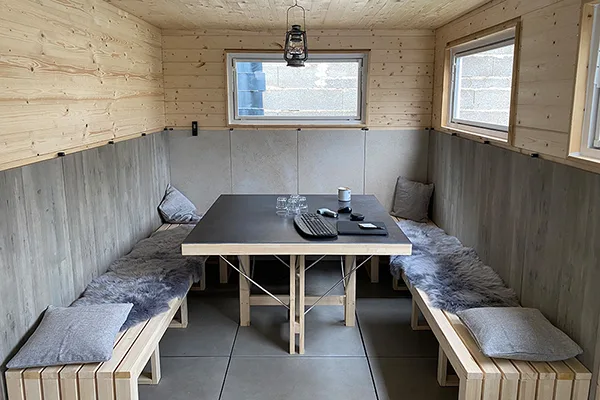 Ein Innenraum, der aus einem umgebauten LKW-Container besteht. Der Raum ist mit Holz verkleidete Wänden, drei große Fenster und eine gemütliche Sitzecke mit integrierten unsichtbaren Infrarotheizelementen. Das Ambiente wirkt warm, einladend und ideal für gesellige Zusammenkünfte