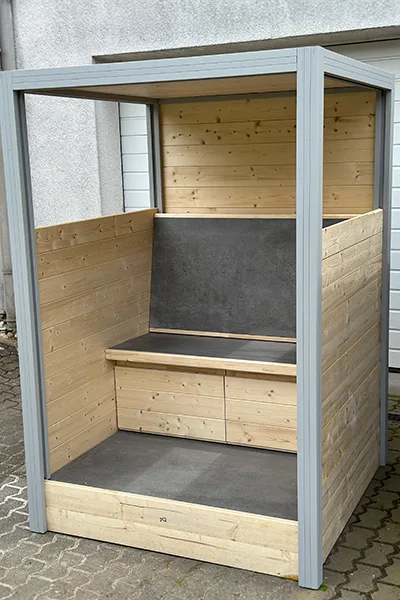 Der eliTile Sitzcube ist eine moderne Sitznische mit einem stabilen Aluminium-Rahmen. Die Konstruktion ist aus Holz mit klaren, minimalistischen Linien gestaltet. Die Seitenwände und die Rückwand bestehen aus hellen Holzpaneelen, während die Sitzfläche, Bodenfläche und Rückenlehne dunkler sind; in ihnen befinden sich die Heizelemente. der eliTile Sitzcube bietet einen geschützten Sitzbereich, der für den Außenbereich geeignet ist.
